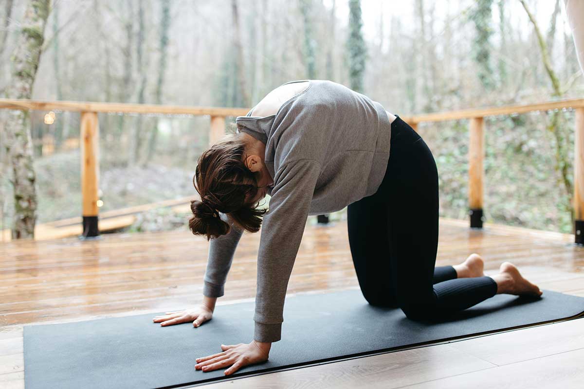 Frau im Vierfüßlerstand kniet auf Yogamatte und macht einen Rundrücken.